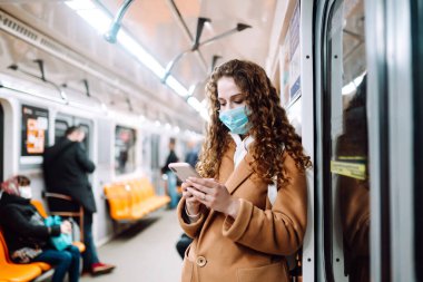 Korunaklı steril tıbbi maskeli bir kız metroda telefonuyla yüzünde. Coronavirus hakkında haber aramak için telefonu kullanan bir kadın. Salgının yayılmasını önleme konsepti.