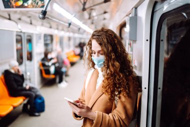 Korunaklı steril tıbbi maskeli bir kız metroda telefonuyla yüzünde. Coronavirus hakkında haber aramak için telefonu kullanan bir kadın. Salgının yayılmasını önleme konsepti.