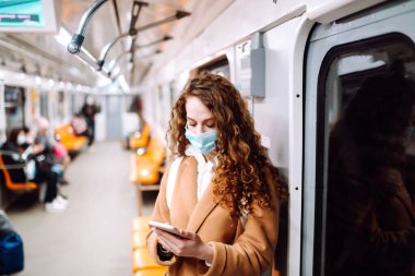 Korunaklı steril tıbbi maskeli bir kız metroda telefonuyla yüzünde. Coronavirus hakkında haber aramak için telefonu kullanan bir kadın. Salgının yayılmasını önleme konsepti.