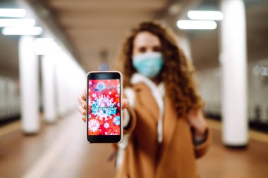 Yüzünde steril tıbbi maske takan kız metro istasyonundaki telefonu uzatıyor. Coronavirus hücreleri telefon ekranında. Salgının yayılmasını önleme konsepti. 
