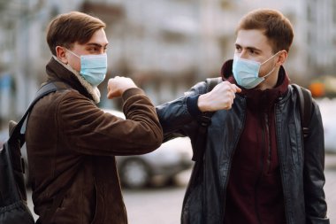 Yüzünde koruyucu tıbbi maske takan arkadaşları dirseklerini karantina şehrinde selamlıyorlar. Dirsek çarpması, koronavirüsün yayılmasını önlemek için yeni bir selamlamadır. El sıkışmayın. El sıkışmayı kesin..
