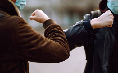 Yüzünde koruyucu tıbbi maske takan arkadaşları dirseklerini karantina şehrinde selamlıyorlar. Dirsek çarpması, koronavirüsün yayılmasını önlemek için yeni bir selamlamadır. El sıkışmayın. El sıkışmayı kesin..