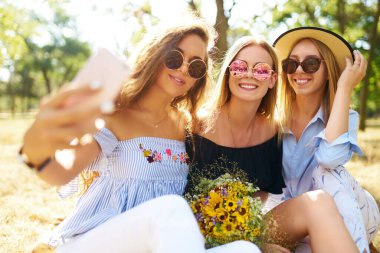 Üç moda genç kız sıcak yaz gününde gülüp selfie çekiyor. Arkadaşlar tatillerinin tadını çıkarır. Birlikte eğlenirler. Yaz konsepti.