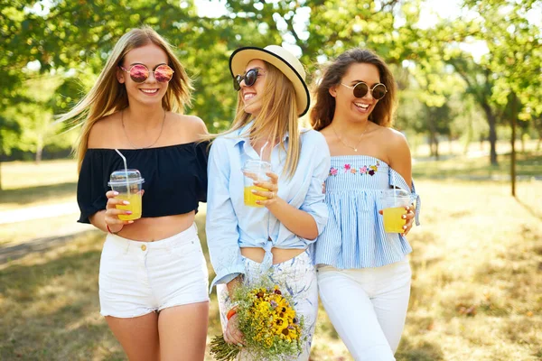 Three beautiful young girls have fun together and drink fresh fruit juice in  hot summer day. Friends enjoying orange juice and  posing against the backdrop of the park. Summer concept.