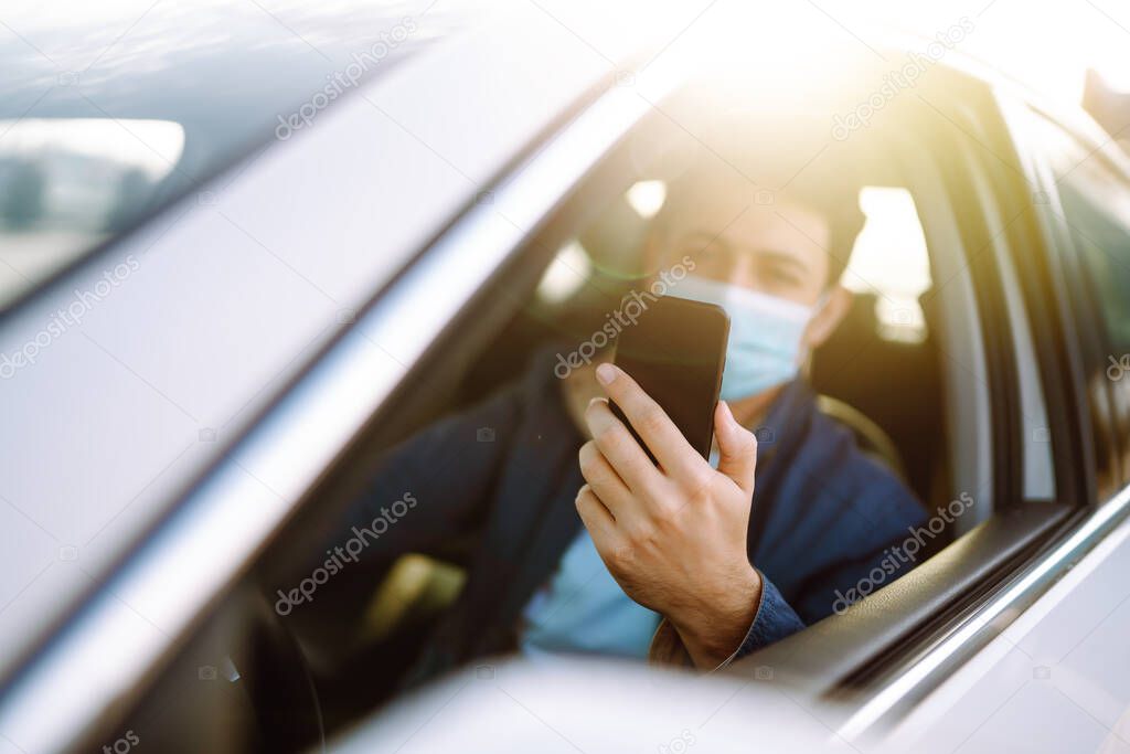 Young man in protective sterile medical mask using phone driving car. The concept of preventing the spread of the epidemic and treating coronavirus, pandemic in quarantine city. Covid -19.