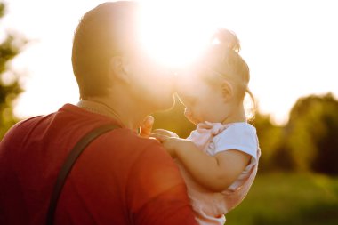 Mutlu baba ve küçük kız gün batımında yaz parkında eğleniyorlar. Babam güneşli arazide küçük bir çocukla oynuyor. Baba ve kızının öpücükleri ve kucaklamaları. Aile ve babalık kavramı.