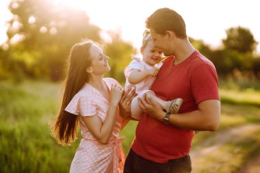 Mutlu genç aile gün batımında parkta yürüyor. Anne, baba ve küçük kız yaz parkında eğleniyorlar. Mutlu aile kavramı. Ebeveynler bebeğin elini tutuyor. Öpücükler ve sarılmalar.