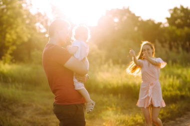 Mutlu genç aile gün batımında parkta yürüyor. Anne, baba ve küçük kız yaz parkında eğleniyorlar. Mutlu aile kavramı. Ebeveynler bebeğin elini tutuyor. Öpücükler ve sarılmalar.