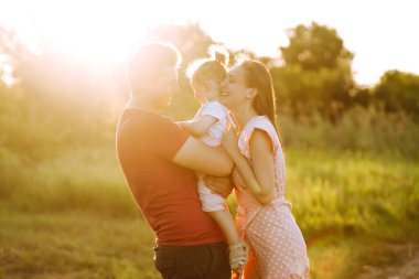 Mutlu genç aile gün batımında parkta yürüyor. Anne, baba ve küçük kız yaz parkında eğleniyorlar. Mutlu aile kavramı. Ebeveynler bebeğin elini tutuyor. Öpücükler ve sarılmalar.