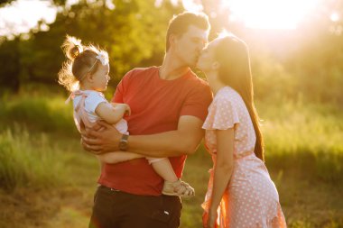 Mutlu genç aile gün batımında parkta yürüyor. Anne, baba ve küçük kız yaz parkında eğleniyorlar. Mutlu aile kavramı. Ebeveynler bebeğin elini tutuyor. Öpücükler ve sarılmalar.
