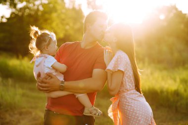 Mutlu genç aile gün batımında parkta yürüyor. Anne, baba ve küçük kız yaz parkında eğleniyorlar. Mutlu aile kavramı. Ebeveynler bebeğin elini tutuyor. Öpücükler ve sarılmalar.
