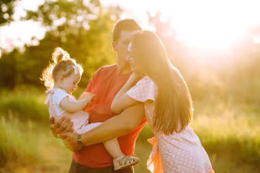 Mutlu genç aile gün batımında parkta yürüyor. Anne, baba ve küçük kız yaz parkında eğleniyorlar. Mutlu aile kavramı. Ebeveynler bebeğin elini tutuyor. Öpücükler ve sarılmalar.
