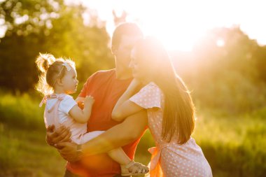 Mutlu genç aile gün batımında parkta yürüyor. Anne, baba ve küçük kız yaz parkında eğleniyorlar. Mutlu aile kavramı. Ebeveynler bebeğin elini tutuyor. Öpücükler ve sarılmalar.