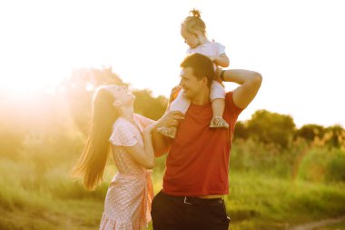 Mutlu genç aile gün batımında parkta yürüyor. Anne, baba ve küçük kız yaz parkında eğleniyorlar. Mutlu aile kavramı. Ebeveynler bebeğin elini tutuyor. Öpücükler ve sarılmalar.