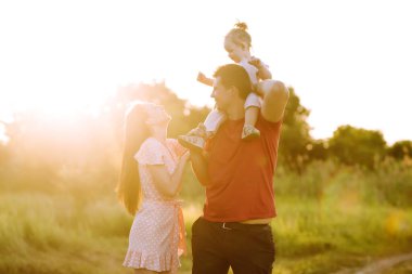Mutlu genç aile gün batımında parkta yürüyor. Anne, baba ve küçük kız yaz parkında eğleniyorlar. Mutlu aile kavramı. Ebeveynler bebeğin elini tutuyor. Öpücükler ve sarılmalar.
