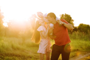 Mutlu genç aile gün batımında parkta yürüyor. Anne, baba ve küçük kız yaz parkında eğleniyorlar. Mutlu aile kavramı. Ebeveynler bebeğin elini tutuyor. Öpücükler ve sarılmalar.