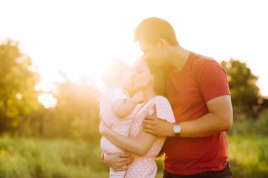 Mutlu genç aile gün batımında parkta yürüyor. Anne, baba ve küçük kız yaz parkında eğleniyorlar. Mutlu aile kavramı. Ebeveynler bebeğin elini tutuyor. Öpücükler ve sarılmalar.