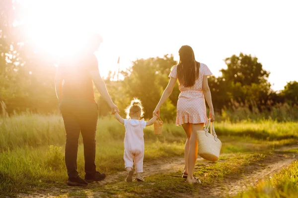 Happy young family walking in the park at sunset. Mom, dad and little daughter having fun in summer park. The concept of a happy family. Parents hold the baby\'s hands. Kisses and hugs.