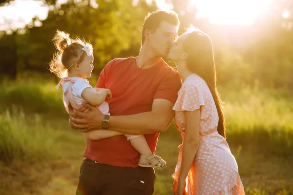 Happy young family walking in the park at sunset. Mom, dad and little daughter having fun in summer park. The concept of a happy family. Parents hold the baby\'s hands. Kisses and hugs.