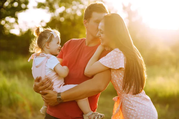 Happy young family walking in the park at sunset. Mom, dad and little daughter having fun in summer park. The concept of a happy family. Parents hold the baby\'s hands. Kisses and hugs.