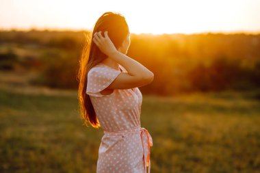 Gün batımında güzel genç bir kadının portresi. Genç kız yaz parkında güneşin tadını çıkarıyor. Mutlu kadın güneşli arazide doğanın tadını çıkarıyor. İnsanlar, yaşam tarzı, rahatlama ve tatil kavramı.