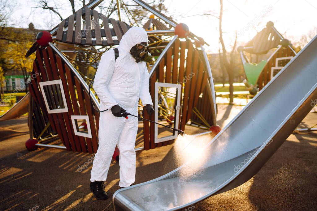 Man wearing protective suit disinfecting the playground in the sun with spray chemicals to preventing the spread of coronavirus, pandemic in quarantine city. Covid -19. Cleaning concept.