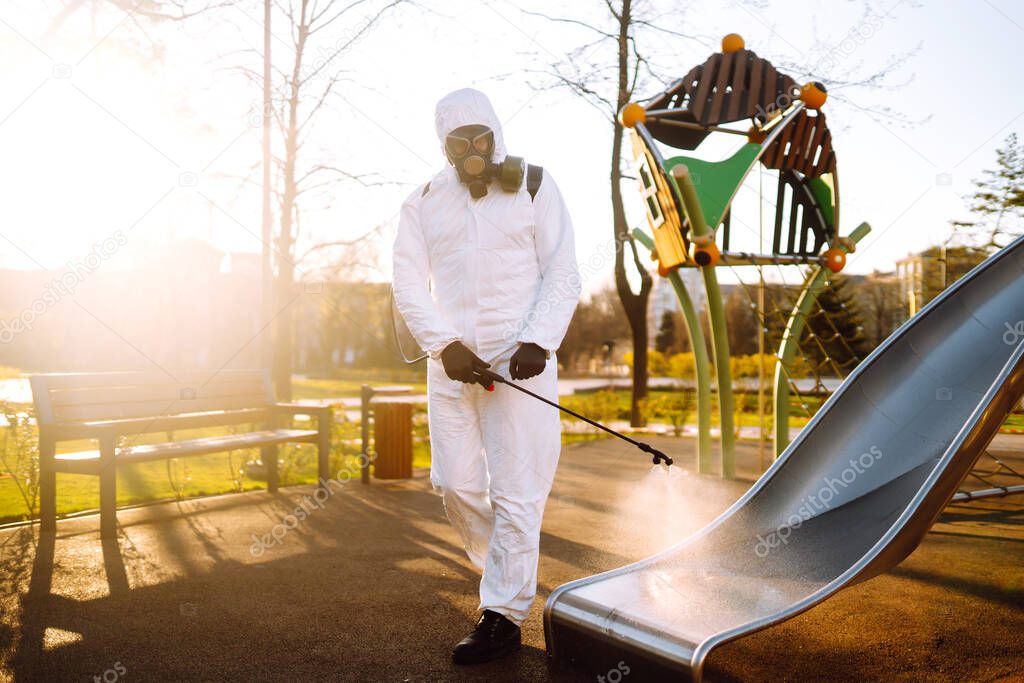 Man wearing protective suit disinfecting the playground in the sun with spray chemicals to preventing the spread of coronavirus, pandemic in quarantine city. Covid -19. Cleaning concept.