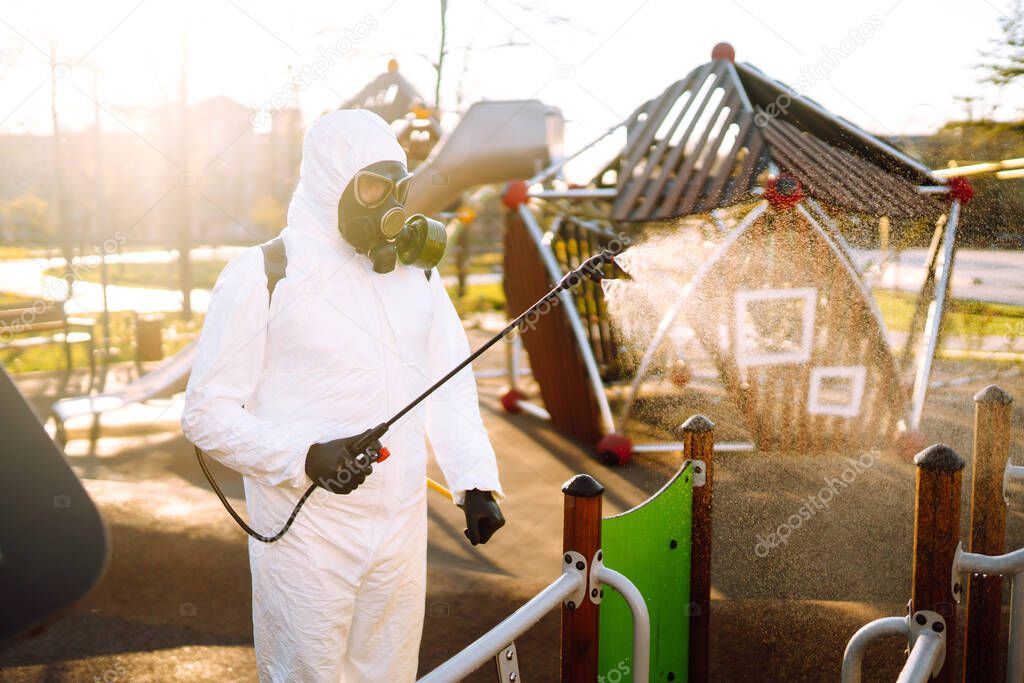 Man wearing protective suit disinfecting the playground in the sun with spray chemicals to preventing the spread of coronavirus, pandemic in quarantine city. Covid -19. Cleaning concept.