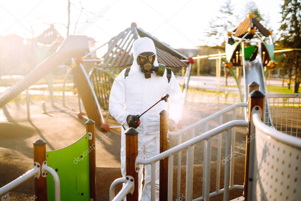 Man wearing protective suit disinfecting the playground in the sun with spray chemicals to preventing the spread of coronavirus, pandemic in quarantine city. Covid -19. Cleaning concept.