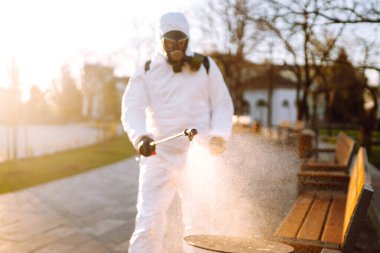 Koruyucu kıyafet giyen bir adam karantina şehrinde salgın olan koronavirüsün yayılmasını önlemek için güneş altındaki halka açık yerleri sprey kimyasallarla dezenfekte ediyor. Covid -19. Temizlik kavramı.