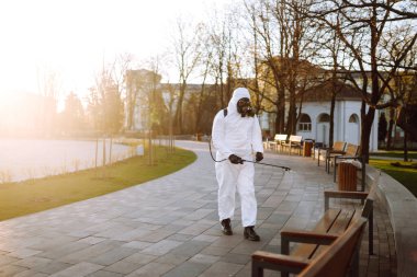 Koruyucu kıyafet giyen bir adam karantina şehrinde salgın olan koronavirüsün yayılmasını önlemek için güneş altındaki halka açık yerleri sprey kimyasallarla dezenfekte ediyor. Covid -19. Temizlik kavramı.