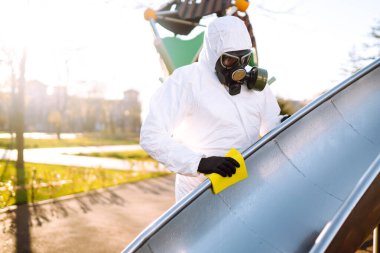 Koruyucu kıyafet giyen adam karantina şehrinde salgın olan koronavirüsün yayılmasını önlemek için oyun alanını temizliyor ve dezenfekte ediyor. Covid -19. Temizlik ve Dezenfeksiyon.