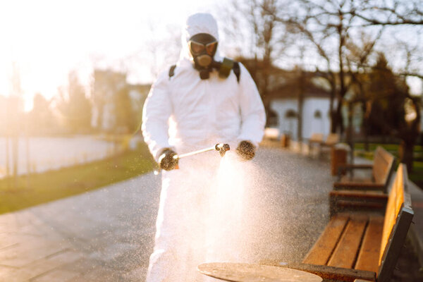 Человек в защитном костюме дезинфицирует общественные места на солнце спреем химикатов для предотвращения распространения коронавируса, пандемии в карантинном городе. Ковид - 19. Концепция очистки
.