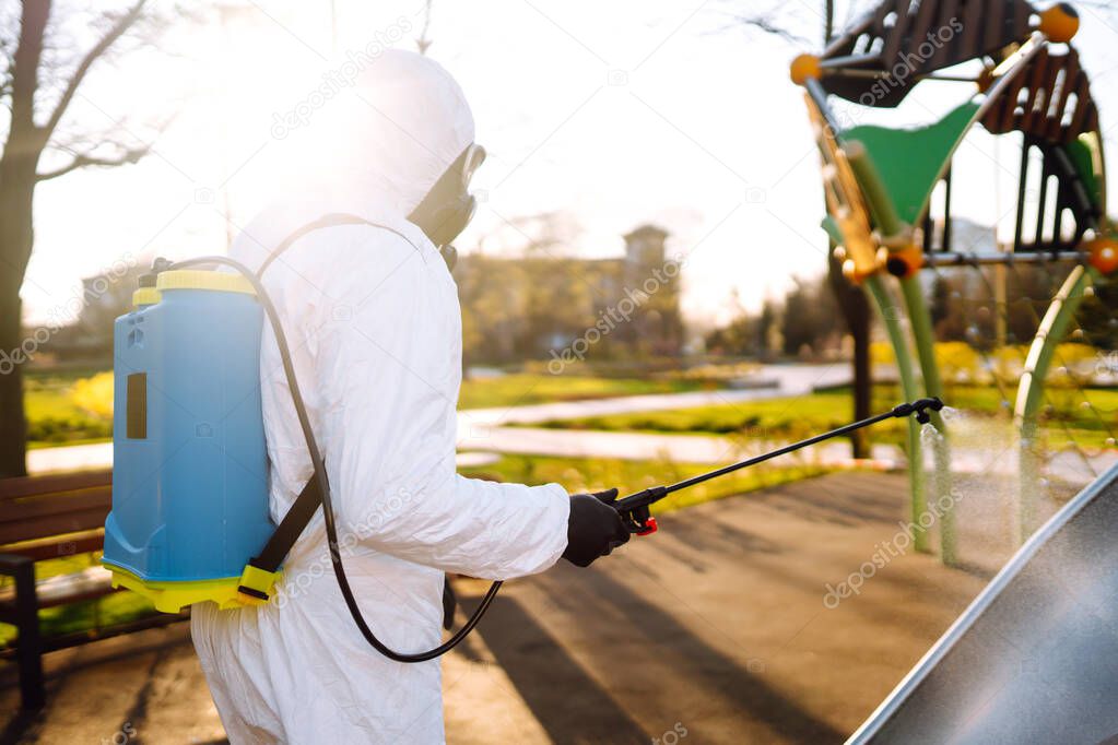 Man wearing protective suit disinfecting the playground in the sun with spray chemicals to preventing the spread of coronavirus, pandemic in quarantine city. Covid -19. Cleaning concept.