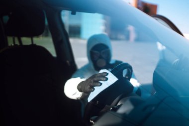 Koruyucu giysili Adam 'ın eli arabadaki direksiyon simidini yıkıyor ve dezenfekte ediyor, Covid-19 virüsü enfeksiyonunu önlüyor, mikrop veya bakteri bulaşmasını önlüyor.