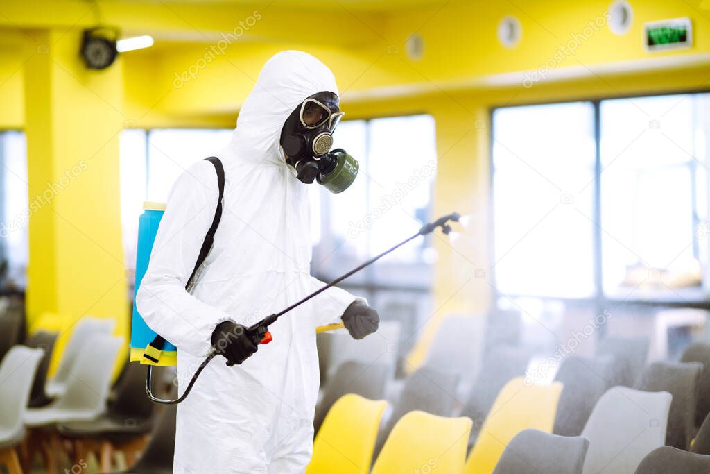 Man wearing protective suit disinfecting assembly hall with spray chemicals to preventing the spread of coronavirus, pandemic in quarantine city. Disinfecting of office. COVID-19.