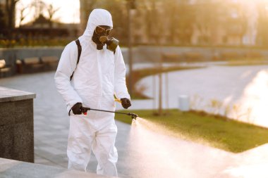 A man wearing special protective disinfection suit sprays sterilizer  in the empty public place at dawn in the city of quarantine. Covid -19. Cleaning concept. clipart