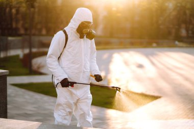 A man wearing special protective disinfection suit sprays sterilizer  in the empty public place at dawn in the city of quarantine. Covid -19. Cleaning concept. clipart