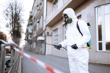 Koruyucu giysili ve maskeli bir adam şafak vakti karantina şehrindeki boş halka açık alanda dezenfektan sıkar. Covid -19. Temizlik kavramı.