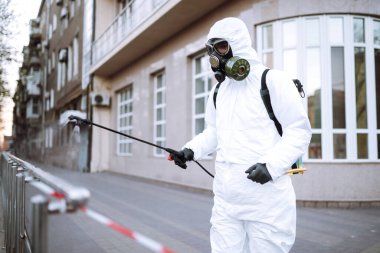 Koruyucu giysili ve maskeli bir adam şafak vakti karantina şehrindeki boş halka açık alanda dezenfektan sıkar. Covid -19. Temizlik kavramı.