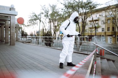 Koruyucu giysili ve maskeli bir adam şafak vakti karantina şehrindeki boş halka açık alanda dezenfektan sıkar. Covid -19. Temizlik kavramı.