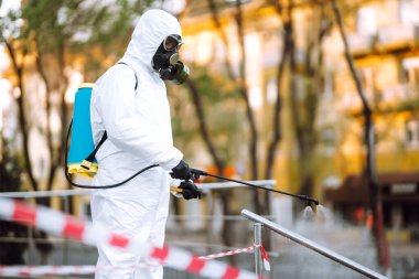 Koruyucu giysili ve maskeli bir adam şafak vakti karantina şehrindeki boş halka açık alanda dezenfektan sıkar. Covid -19. Temizlik kavramı.