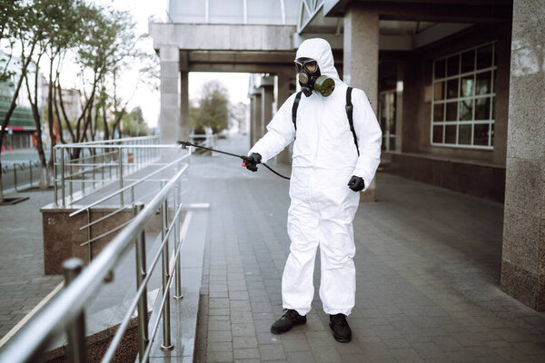 Man in protective suit  and mask sprays disinfector onto the railing in the empty public place at dawn in the city of quarantine. Covid -19. Cleaning concept.