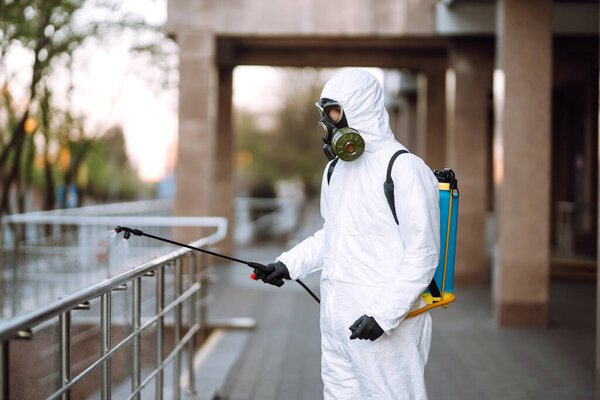 Человек в защитном костюме и маске дезинфицирует перила в пустом общественном месте на рассвете в городе карантина. Ковид - 19. Концепция очистки
.