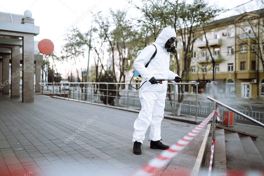 Man in protective suit  and mask sprays disinfector onto the railing in the empty public place at dawn in the city of quarantine. Covid -19. Cleaning concept.