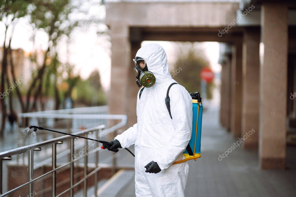 Man in protective suit  and mask sprays disinfector onto the railing in the empty public place at dawn in the city of quarantine. Covid -19. Cleaning concept.