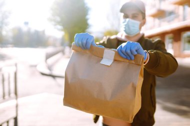 Koruyucu maskeli ve tıbbi eldivenli kurye, içinde yiyecek olan kağıt paketler tutuyor. Karantina şehrinde bir scooterla teslimat servisi. Covid 19.
