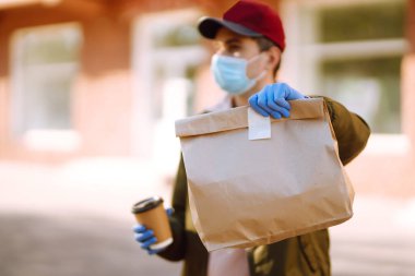 Koruyucu maskeli ve tıbbi eldivenli kurye, içinde yiyecek olan kağıt paketler tutuyor. Karantina şehrinde bir scooterla teslimat servisi. Covid 19.