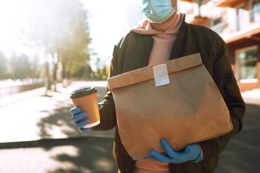 Koruyucu maskeli ve tıbbi eldivenli kurye, içinde yiyecek olan kağıt paketler tutuyor. Karantina şehrinde bir scooterla teslimat servisi. Covid 19.