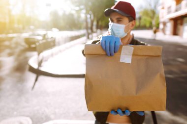 Koruyucu maskeli ve tıbbi eldivenli kurye, içinde yiyecek olan kağıt paketler tutuyor. Karantina şehrinde bir scooterla teslimat servisi. Covid 19.